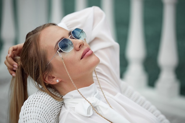 Porträt einer Blondine mit Sonnenbrille, die auf einem Stuhl an der frischen Luft ruht