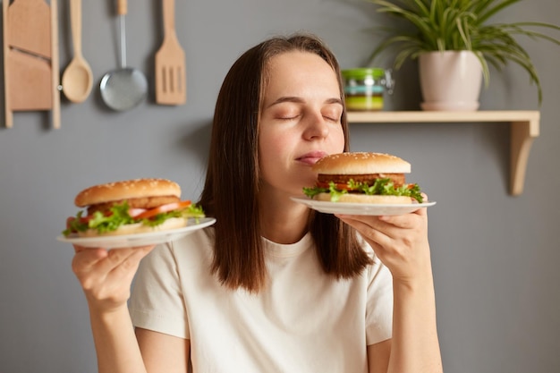 Porträt einer braunhaarigen kaukasischen Frau, die mit geschlossenen Augen und zufriedenem Gesichtsausdruck zwei köstliche frische Hamburger hält, die nach Fast Food duften