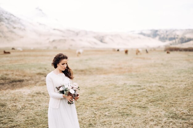 Porträt einer Braut in einem weißen Hochzeitskleid mit einem Brautstrauß in den Händen auf einem trockenen Feld