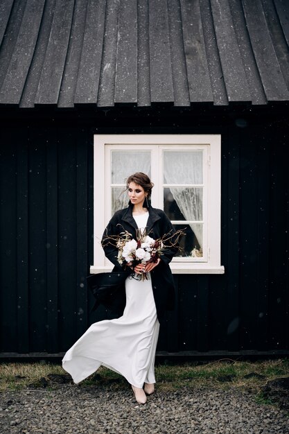Porträt einer Braut in einem weißen Seidenhochzeitskleid und einem schwarzen Mantel mit einem Brautstrauß