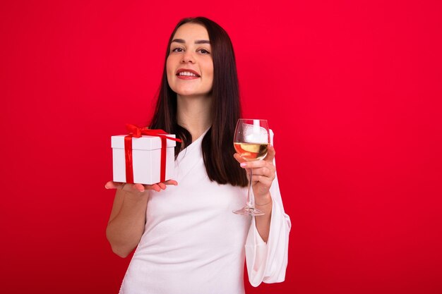 Porträt einer Brünette in einem weißen Kleid mit einem Glas Wein und einer Geschenkbox auf rotem Hintergrund Eine junge Frau feiert das neue Jahr