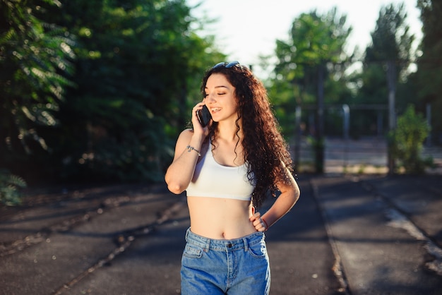 Porträt einer Brünette mit langen lockigen Haaren, die in einem weißen Oberteil und Jeans auf der Straße steht und am Telefon spricht. Ein lustiges Gespräch.