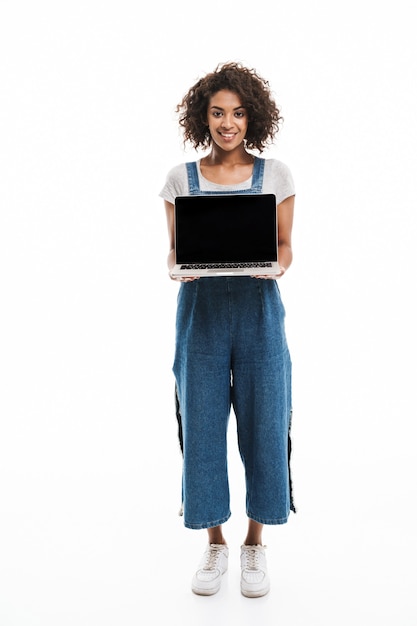 Porträt einer brünetten Frau, die in Denim-Overalls gekleidet ist und den offenen Laptop isoliert über der weißen Wand hält?