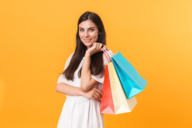 Porträt einer brünetten Shopaholic-Frau, die ein Kleid trägt, das lächelt, während sie bunte Einkaufstüten isoliert über gelber Wand hält?