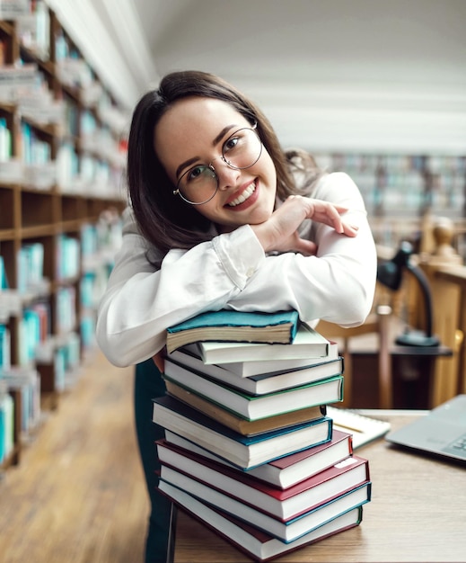 Porträt einer brünetten Teenagerin, die Bücher in der Nähe des Tisches in der Bibliothek hält