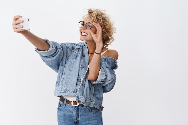 Porträt einer charmanten, enthusiastischen und stilvollen, gut aussehenden blonden Frau in Jeansjacke und Brille, die breit auf das Smartphone lächelt, während sie ein Selfie auf der Frontkamera macht