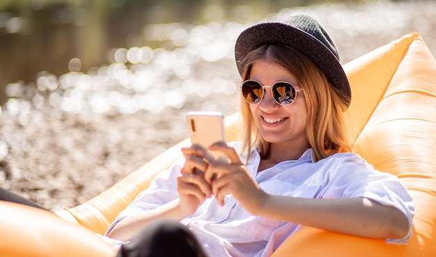 Porträt einer charmanten lächelnden Frau mit Sonnenbrille, die ein Handy benutzt und Nachrichten an Freunde schreibt, während sie auf einer Luftmatratze am Sommerstrand liegt