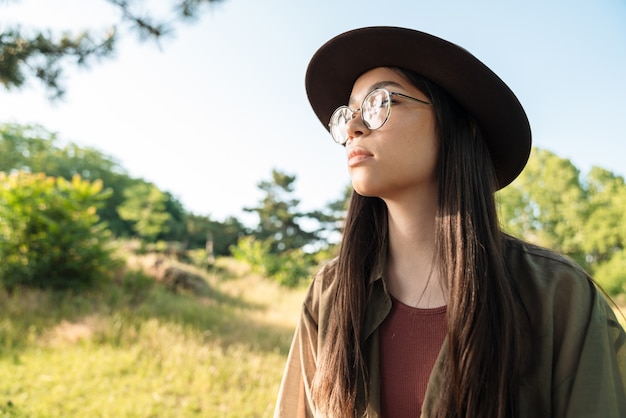 Porträt einer denkenden jungen Frau mit langen dunklen Haaren, die einen stilvollen Hut und eine Brille trägt, die an einem sonnigen Tag im grünen Park spazieren gehen?