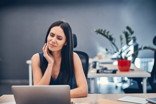 Porträt einer desillusionierten Dame, die beim Arbeiten mit Laptop im Büro Schmerzen im Zahn verspürt