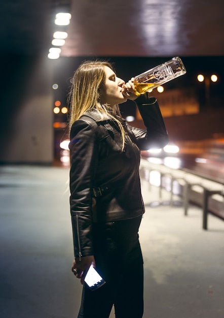 Porträt einer einsamen Frau, die Alkohol aus der Flasche an der Autobahn trinkt