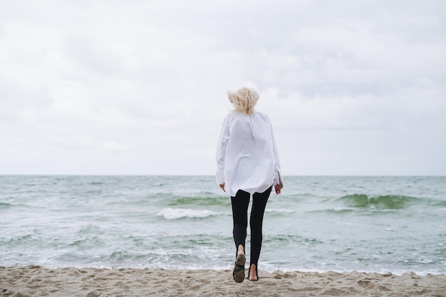 Porträt einer eleganten blonden Frau in weißem Hemd am Sandstrand bei Sturmmeer bei windigem Wetter
