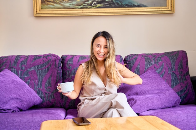 Porträt einer eleganten jungen Frau, die zu Hause mit einem Kaffee in der Hand auf dem Sofa sitzt