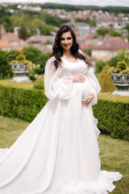 Porträt einer eleganten schwangeren Frau in schönem weißem Kleid, die im Freien spazieren geht