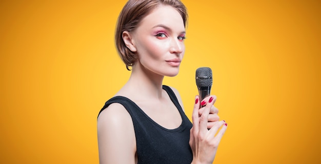 Foto porträt einer eleganten, stilvollen frau mit mikrofon. gelber hintergrund. karaoke-konzept. gemischte medien