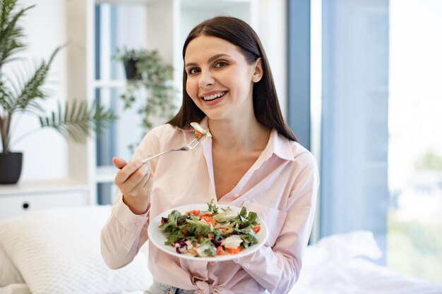 Porträt einer entspannten Frau, die auf einem bequemen Bett sitzt und einen gesunden Salat isst