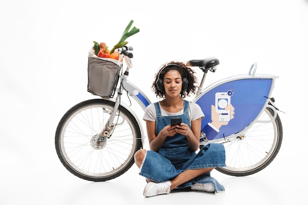 Porträt einer entspannten Frau, die Handy hält, während sie in der Nähe des Fahrrads sitzt, mit Einkaufstasche isoliert über weißer Wand