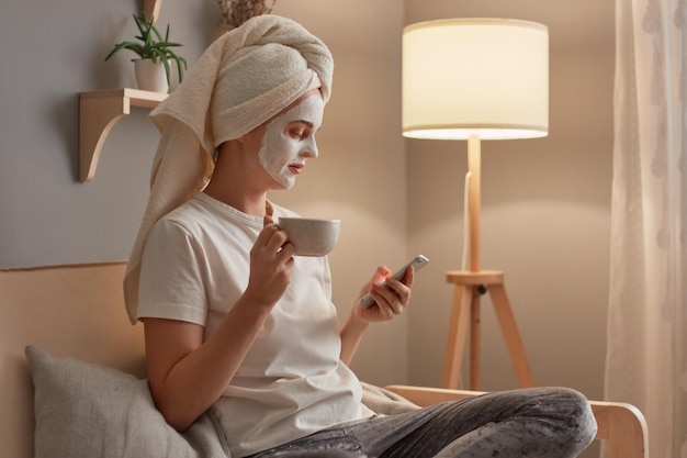 Porträt einer entspannten Frau in weißem T-Shirt und Handtuch auf dem Kopf, die auf Husten sitzt und mit Schönheitsbehandlungsmaske auf ihrem Gesicht posiert, Kaffee nippt und mit ihrem Telefon soziale Netzwerke überprüft