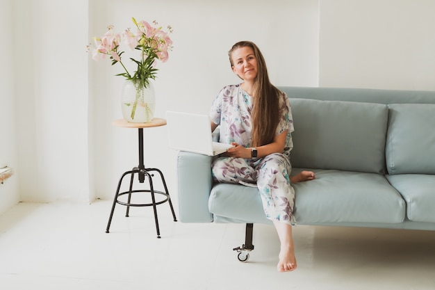 Porträt einer entzückenden jungen Frau mit Laptop, die zu Hause arbeitet, während sie auf einem blauen Sofa in einem gemütlichen Zimmer sitzt