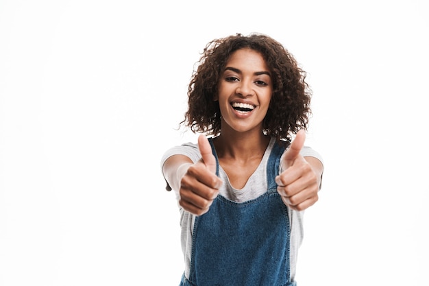 Porträt einer entzückten Frau, die in Denim-Overalls gekleidet ist und vorne mit Daumen nach oben lächelt, isoliert über weißer Wand?