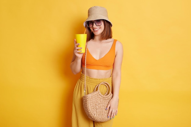 Porträt einer entzückten jungen dunkelhaarigen Frau mit Sonnenbrille in Sommerkleidung, die isoliert vor gelbem Hintergrund posiert und eine Tasse mit leckerem frischem Getränk hält, die lächelnd in die Kamera schaut