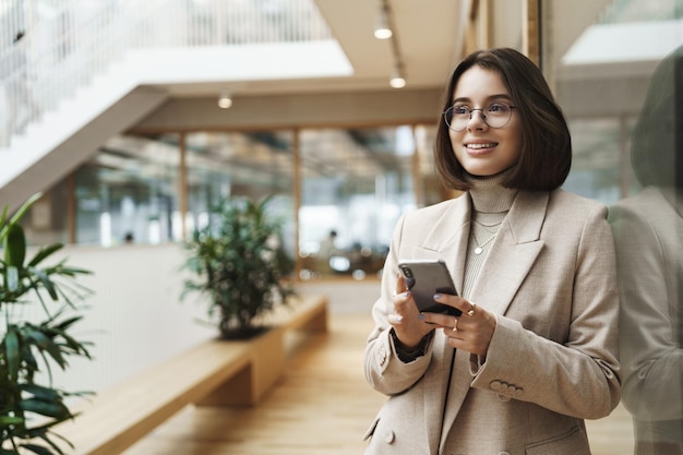 Porträt einer erfolgreichen, glücklich lächelnden Frau, die gerade ein Treffen mit Geschäftspartnern abgeschlossen hat, unterzeichnete ein Geschäft, das ein Mobiltelefon hält und mit einem verträumten, zufriedenen Ausdruck wegschaut