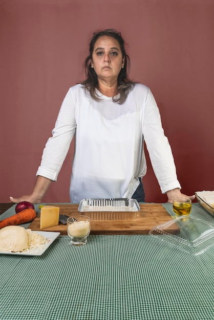 Porträt einer ernsthaften Frau, die vor verschiedenen Produkten für die Zubereitung eines Lasagne-Rezeptes in die Kamera blickt