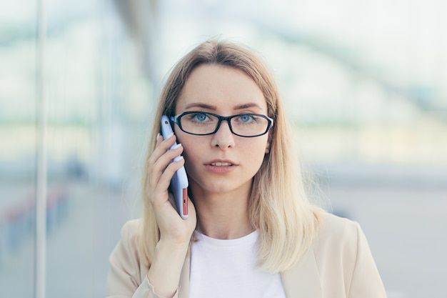 Porträt einer ernsthaften Frau mit Brille blond, die ein Mobiltelefon hält und auf einem Smartphone in der Nähe des Büros spricht