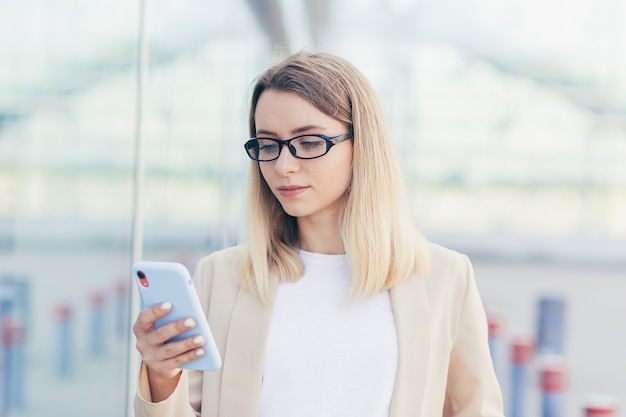 Porträt einer ernsthaften Geschäftsfrau mit blonder Brille liest die Nachrichten von einem Mobiltelefon und verwendet die Anwendung, um Nachrichten umzuschreiben