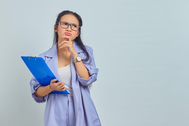 Porträt einer ernsthaften jungen asiatischen Geschäftsfrau, die das Kinn hält und über etwas nachdenkt, während sie den Dokumentenordner isoliert auf violettem Hintergrund hält