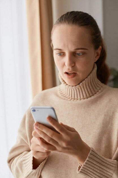 Porträt einer erstaunten, erstaunten Frau mit Pferdeschwanzfrisur, die einen beigefarbenen Pullover im lässigen Stil trägt, der mit einem Handy in den Händen steht und mit schockiertem Gesichtsausdruck auf das Gadget-Display blickt.