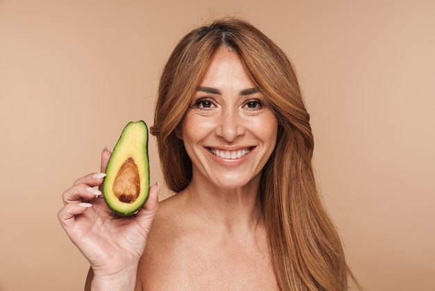 Porträt einer erwachsenen Frau mit nacktem Oberkörper, die Avocado hält und isoliert auf Beige lächelt