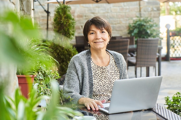 Porträt einer erwachsenen Geschäftsfrau mit Laptop am Tisch im Sommercafé, die draußen arbeitet und in die Kamera lächelt Selbstständiges Konzept für freiberufliche ältere Frauen