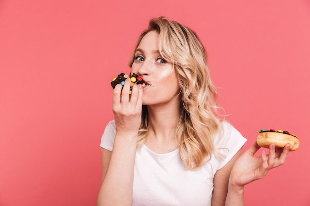 Porträt einer europäischen blonden Frau, die ein lässiges T-Shirt trägt, das leckere süße Donuts hält und isst, die über rosa Wand isoliert sind?