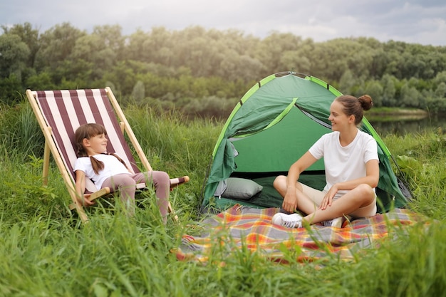 Porträt einer Familie, die in der Nähe des Flusses reist und campt, sitzt in einem Klappsessel und einer Decke auf dem Boden und entspannt sich am Seeufer und fühlt sich gemeinsam glücklich