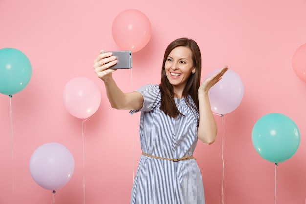 Porträt einer faszinierenden lächelnden Frau in blauem Kleid, die Selfie auf dem Handy macht und die Hände auf rosafarbenem Hintergrund mit bunten Luftballons ausbreitet. Geburtstagsfeier, Konzept der aufrichtigen Emotionen der Menschen.