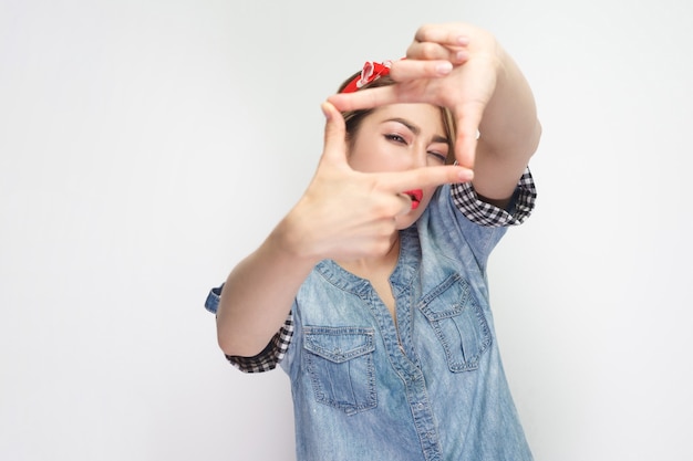 Porträt einer fokussierten schönen jungen Frau im lässigen blauen Jeanshemd mit rotem Stirnband, das mit Erntekompositionsgeste steht und Kamera betrachtet. Indoor-Studioaufnahme, isoliert auf weißem Hintergrund.