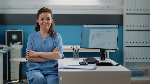 Porträt einer Frau, die als Arzthelferin am Schreibtisch in der Arztpraxis arbeitet. Krankenschwester schaut in die Kamera und lächelt, während sie sich auf die Arbeit am Computer vorbereitet. Gesundheitsspezialist in Uniform