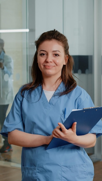 Porträt einer Frau, die als Krankenschwester arbeitet und Kontrollpapiere hält, während sie in der Krankenstation steht. Arzthelferin mit Dokumenten, die die Kamera betrachten und lächeln und dem Spezialisten helfen.