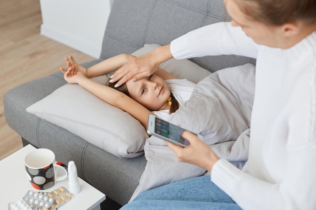 Porträt einer Frau, die auf dem Sofa neben der kranken Tochter sitzt, die Hand auf die Stirn hält und mit dem Smartphone in den Händen posiert und den Arzt anruft, um nach der Behandlung zu fragen