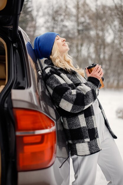 Porträt einer Frau, die im Winterwald in der Nähe des Kofferraums eines offenen Autos steht