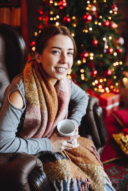 Porträt einer Frau, die in einem Sessel neben dem Weihnachtsbaum mit rotem Spielzeug sitzt und eine Tasse in ihren Händen hält