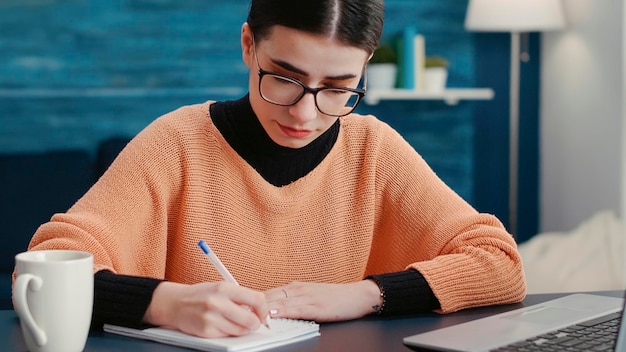 Porträt einer Frau, die lächelt und einen Kursbericht auf einem Notizbuch schreibt und mit einem Laptop am Schreibtisch sitzt. Online-Schüler, der sich Notizen zum Lehrbuch macht und Remote-Unterrichtssitzungen zuhört.