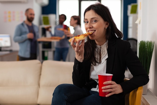 Porträt einer Frau, die nach Feierabend bei einer Partyfeier im Geschäftsbüro Pizza isst. Fröhlicher Arbeiter, der sich beim Treffen mit Getränken, Essen, Spaß und Freizeitaktivitäten entspannt fühlt.