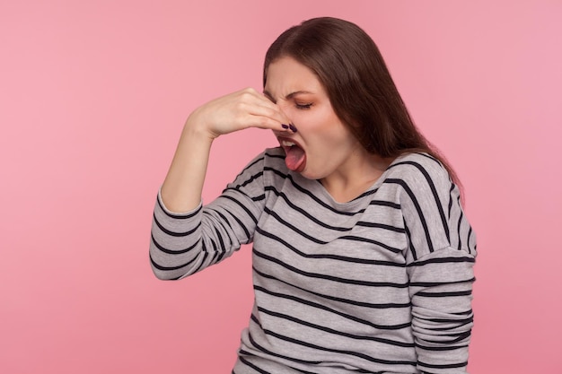 Porträt einer Frau, die vor Ekel das Gesicht verzieht und die Nase mit den Fingern in die Nase drückt, um einen schlechten Geruch zu vermeiden, der die Abneigung zeigt, einen schrecklichen Geruch zu riechen, Studioaufnahme isoliert auf rosa Hintergrund