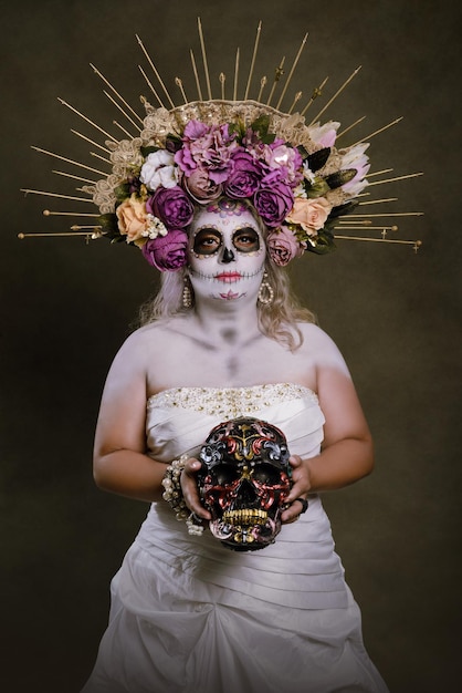 Porträt einer Frau in Catrina-Make-up, die ein Hochzeitskleid trägt, das einen bunt bemalten Schädel hält