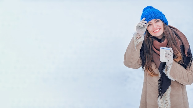 Porträt einer Frau in einem blauen Hut mit einer Tasse auf einem Hintergrund des Schnees