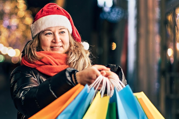 Foto porträt einer frau in einem weihnachtsmannshut mit weihnachtsgeschäften in bunten taschen auf der geschmückten straße