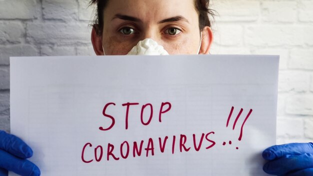 Foto porträt einer frau in einer schutzmaske, die die kamera zeigt, ein weißes blatt papier mit dem text stopp coronavirus in roter markierung. das konzept des quarantäne-coronavirus in der welt.