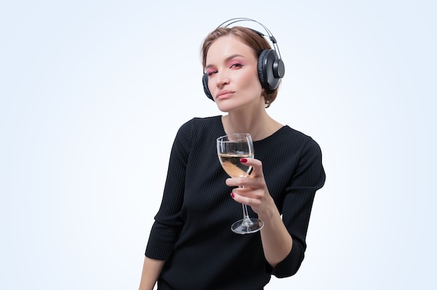 Porträt einer Frau in professionellen Kopfhörern mit einem Glas Wein in der Hand. Weißer Hintergrund. DJ-Konzept. Gemischte Medien