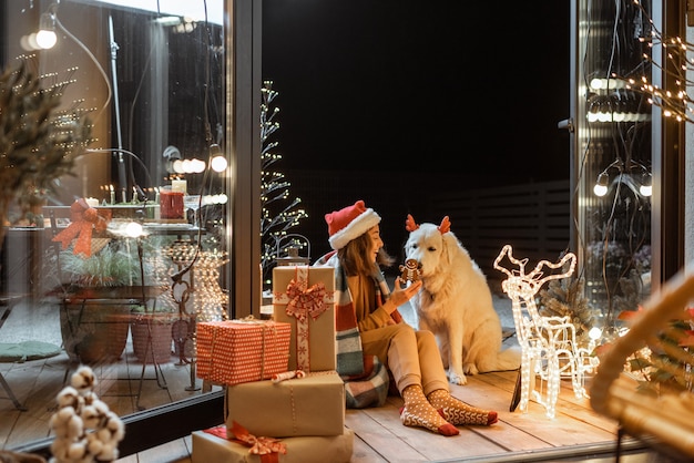 Porträt einer Frau in Weihnachtsmütze und Plaid mit ihrem süßen Hund, der auf der wunderschön dekorierten Terrasse zu Hause einen Neujahrsurlaub feiert und den Hund mit Lebkuchen füttert